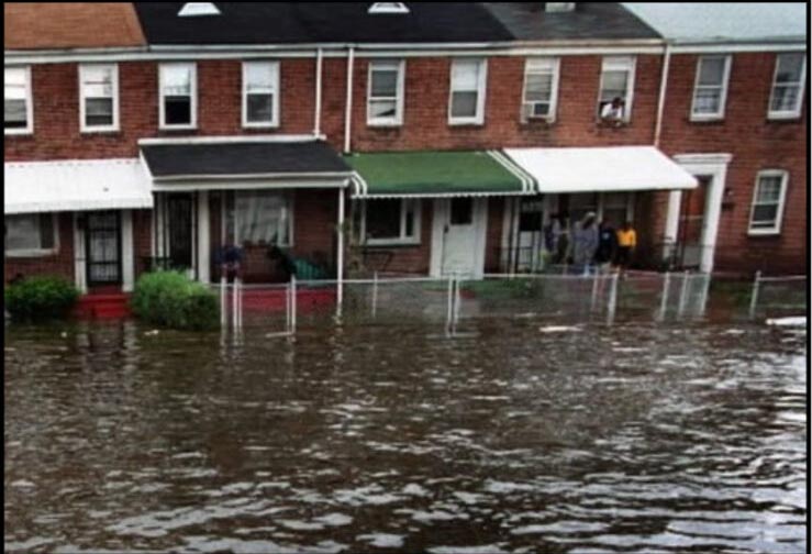 A flooded street in Turner Station, Maryland. The image shows a residential area with water covering the road and reaching up to the sidewalks. Houses and trees are visible in the background, and the sky is overcast.